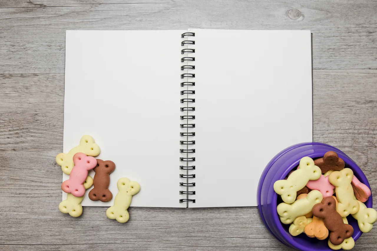 Caderno espiral aberto e em branco sobre uma mesa de madeira, ao lado de biscoitos em formato de osso e uma tigela cheia deles.